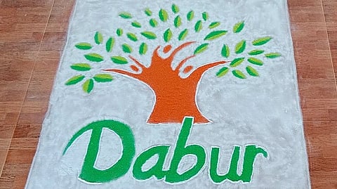 A textured rangoli design on a brown tiled floor features an orange tree with green leaves. Below, the word "Dabur" appears in green, highlighting nature and vibrancy.