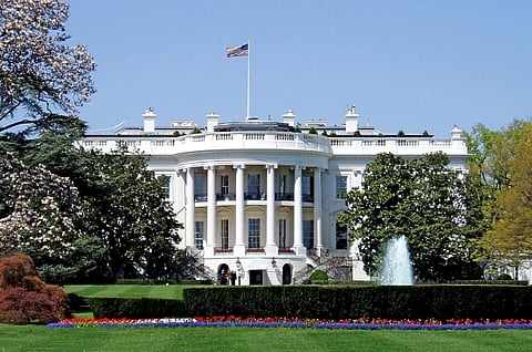 Image of the white house's south facade with its distinctive columns, an American flag atop, surrounded by lush gardens, trees, and a central fountain.