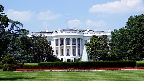 The White House, a grand neoclassical building, is seen behind a manicured lawn and vibrant garden with a central fountain, under a bright blue sky.
