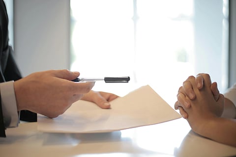 A person in a suit extending a pen and paper across a table to another person with clasped hands. The setting suggests a formal or business context.