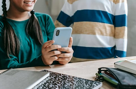 Two children at a desk are smiling while looking at a smartphone. Notebooks and a pencil case are nearby, creating a relaxed learning atmosphere.