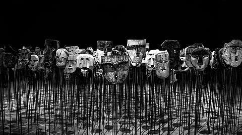 A black and white image of numerous aged, stone-like masks mounted on tall, slender rods. The shadows form an eerie atmosphere on the patterned floor.