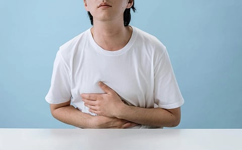 A person in a white T-shirt sits at a table against a blue background, clutching their stomach with a pained expression, conveying discomfort.