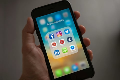 A hand holds a smartphone displaying a folder labeled "Social Networks" with apps like Facebook, Instagram, Twitter, and LinkedIn on the screen.