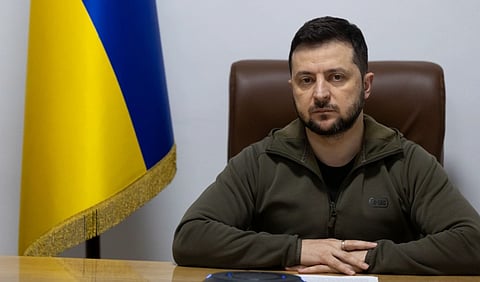 President Zelenskyy in the Romanian Parliament. He is wearing a green zip-up jacket, appearing serious. A Ukrainian flag is visible in the background.
