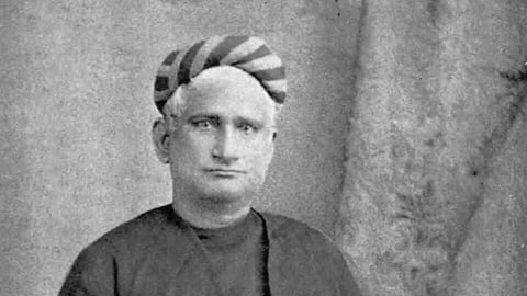 Bankin Chandra Chatterjee stands with a neutral expression, wearing a long dark robe and a striped turban. He holds a book and rests a hand on a chair, evoking a scholarly tone.