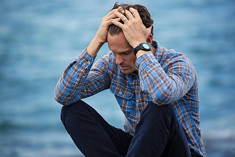 A person in a plaid shirt sits by the water, head in hands, appearing distressed. The ocean backdrop adds a somber, reflective tone to the scene.