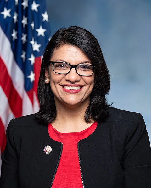 This image depicts a portrait of US Lawmaker Rashida Tlaib, formally dressed with an American flag in the background.