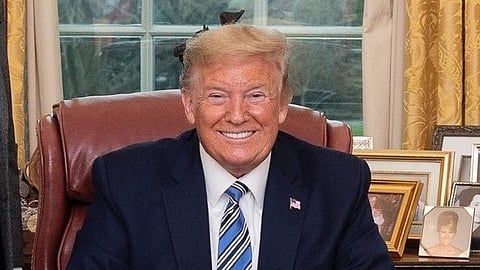 Donald Trump in a suit seated at a a desk. An American flag and framed photos are in the background.