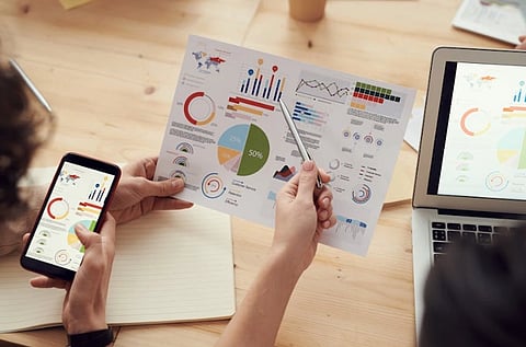  Two people review colorful charts on a paper, phone, and laptop. The setting is a wooden table, creating a collaborative and analytical tone.