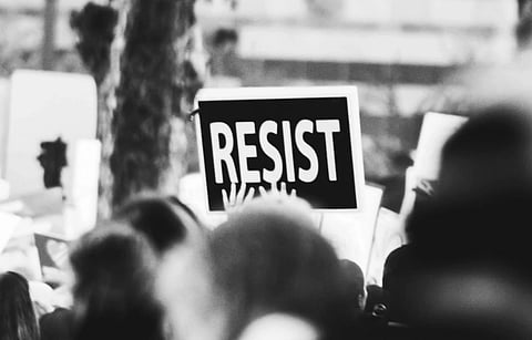 Black-and-white photo of a protest with a prominent sign reading "RESIST." Crowd in the foreground, atmosphere tense and determined.