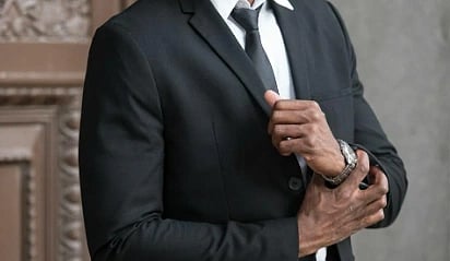 Man in a black suit and tie stands confidently, adjusting his jacket. The ornate brown door in the background adds elegance to the scene.