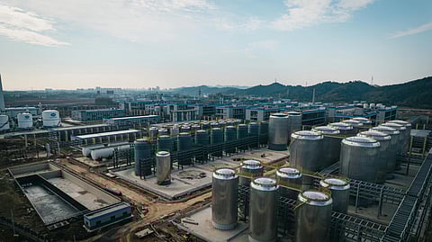 Aerial view of an industrial complex with numerous large, silver storage tanks and buildings, set against distant hills and a cloudy blue sky in China.