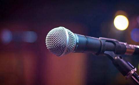Close-up of a microphone on a stand, set against a blurred background with warm, colorful lights. The scene conveys anticipation and focus.