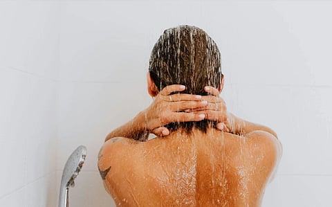 Rear view of a person showering, with water cascading down their back. They have their hands clasped behind their head, creating a calm, relaxing atmosphere.
