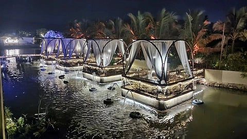 Elegant outdoor dining pods float on a serene pond at night, surrounded by palm trees. Soft lighting and flowing drapes create a romantic ambiance.