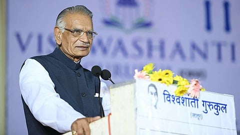 Former Union Home Minister Shivraj Patil in a white shirt and dark vest speaks at a podium adorned with flowers. Behind him is a blurred backdrop with the word "VISWASHANTI."