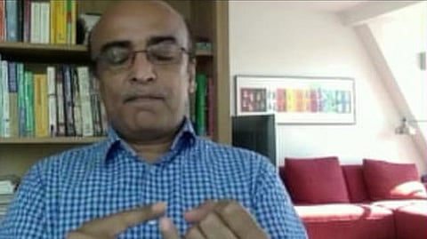 In the image shafi Burfat is shown talking about something. Behind him is book shelf full of books