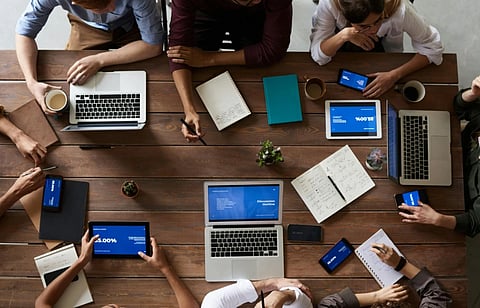 A group of people sit around a wooden table with laptops, tablets, and phones displaying charts. Notebooks and coffee cups are also on the table, indicating a collaborative meeting.