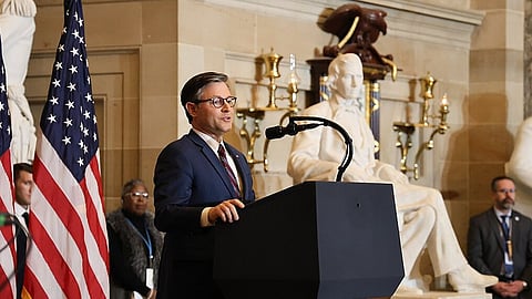 Mike Johnson in a suit speaks confidently at a podium with a microphone. Behind him are two American flags and large statues. The atmosphere is formal and official.