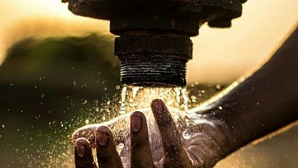 A hand cupped under a rusty outdoor faucet catches a stream of water. Sunlight highlights the splashing droplets, creating a serene and hopeful tone.