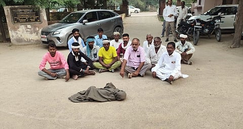 A group of men sit on the road in front of the covered body of a child in protest. Cars and bikes are visible in the background.