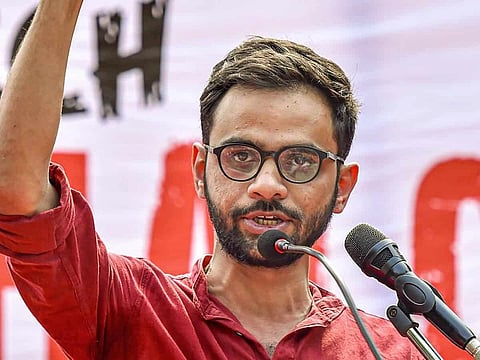 Umar Khalid is shown in this image, with his arm raised speaking at a university campus. He is wearing red coloured shirt, and round framed spectacles.