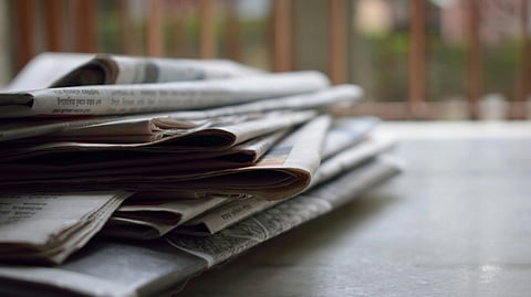 Stack of newspapers on a smooth surface, with a blurred background of vertical bars and greenery.