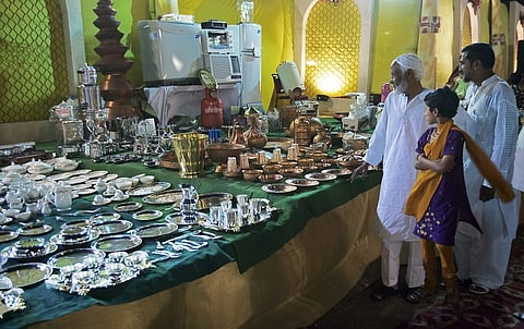 An old man, a middle-aged man, and a young girl stand in front of a clothed table full of silverware and brass utensils.