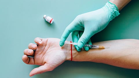 A gloved hand holds a syringe near an outstretched forearm with a red line. A small vaccine vial is nearby on the green background. 
