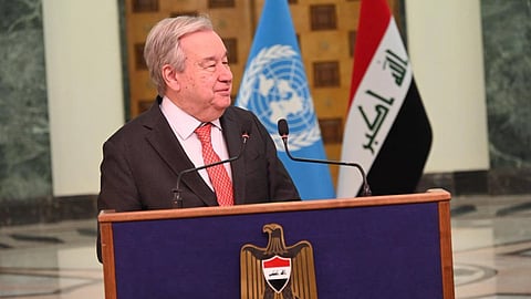 UN Secretary-General Antonio Guterres in a suit speaks at a podium with blue UN and Iraqi flags behind him, conveying a formal setting. Emblem on the podium enhances the official tone.