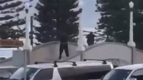 CCTV Footage of two men dressed in black with guns standing on a white arched structure with a parking lot with several cars in the foreground. 