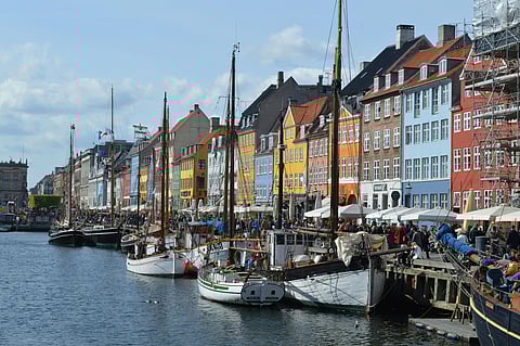 Colorful buildings line a canal with docked sailboats in front. People walk along the vibrant waterfront, creating a lively, bustling atmosphere.