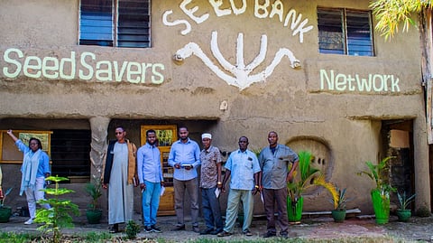 Seven people stand smiling in front of a building of the NGO "Seed Bank Seed Savers Network" with green plants in the foreground.