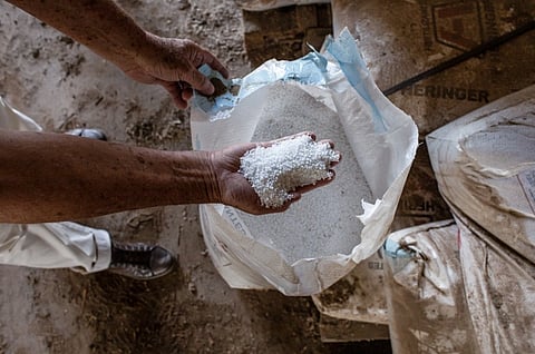 A person holds a handful of granular white fertilizer from an open bag, surrounded by more bags on a dusty floor, conveying a sense of agricultural work.