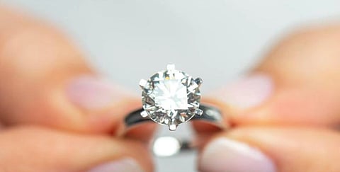 A close-up of hands holding a silver engagement ring with a large, sparkling diamond. The background is blurred, emphasizing the ring's brilliance.
