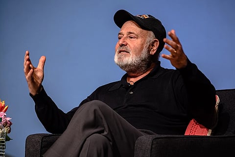 Rob Reiner is stiitng on a couch with legs folded and arms wide, as if explaining something. He is wearing a black cap and a black t-shirt.