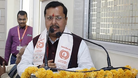 Bihar Minister Nitin Nabin speaking  at a podium decorated with yellow flowers, wearing traditional attire with a red mark on his forehead. He conveys a solemn tone. Two men are visible in the background.