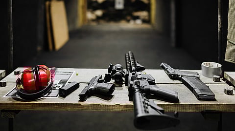 A shooting range table displays firearms, a red earmuff for hearing protection, magazines, and ammunition. The background shows a blurred target area.