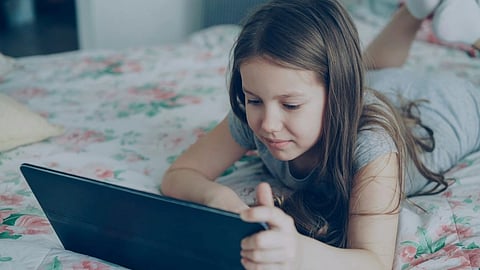 A young girl using a tablet while lying in bed, on floral sheets.