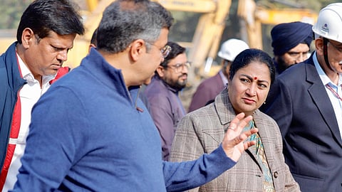 A group of people, including Rekha Gupta in a plaid coat and and Health Minister Pankaj in a blue sweater, walk together at a construction site, conveying a focused and engaged atmosphere.