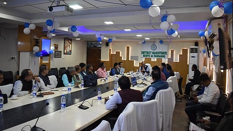 A conference room with DM and DEO conducting a meeting in a U-shaped table. The room is decorated with blue and white balloons with a sign reading "District Magistrate North 24 Parganas" at the front.