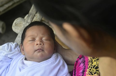 Close-up of a sleeping baby in white clothing held by an adult. The scene conveys warmth and tenderness, with a soft, serene atmosphere.