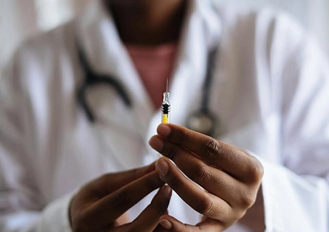 A healthcare professional in a white coat, holding a syringe with yellow liquid. The focus is on steady hands and the syringe; the stethoscope is visible.