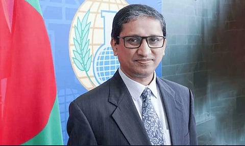  M Riaz Hamidullah in a suit and glasses stands in front of a blue backdrop with a globe and wheat emblem. A red and green flag is visible to the left. He smiles slightly.