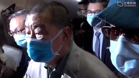 A group of people including Jimmy Lai wearing blue face masks are seen in close quarters. The central figure is in focus, wearing a light suit, conveying a tense atmosphere.