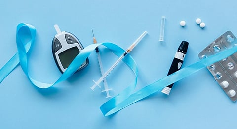 A flat lay of diabetes management tools on a blue background includes a glucometer, insulin syringes, pills, an injection pen, and a blue awareness ribbon.