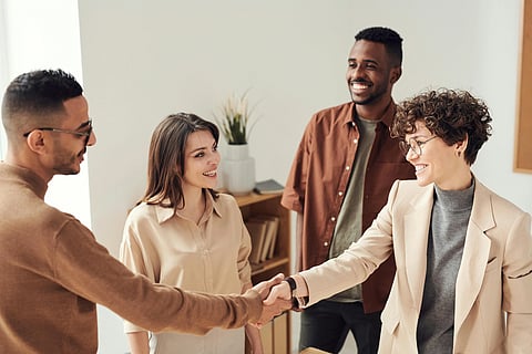 Four diverse colleagues in an office setting are smiling. Two are shaking hands, conveying a sense of agreement and friendly cooperation.