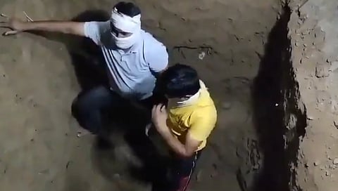 Two people with cloths covering their eyes are inside a dirt pit. One wears a gray shirt, the other a yellow shirt, creating a tense atmosphere.