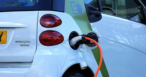 Close-up of a white electric car being charged, with an orange cable connected to the charging port. The car's rear features the text "electric drive."